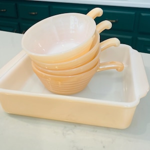 May include: A stack of four peach-colored, ribbed bowls with handles, resting on a matching rectangular baking dish. The bowls and dish have a glossy finish, and are likely made of glass or ceramic. The set is displayed on a white countertop.