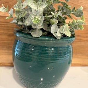May include: A dark teal ceramic planter with a glossy finish, featuring horizontal ridges around the upper body and a textured pattern near the base. The planter contains a faux eucalyptus plant with light green, rounded, dusty-looking leaves.