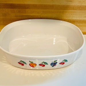 May include: White oval serving dish with a fruit design. The dish has a smooth, glossy surface and a decorative border featuring illustrations of cherries, an orange, and blueberries. The dish is suitable for serving food.