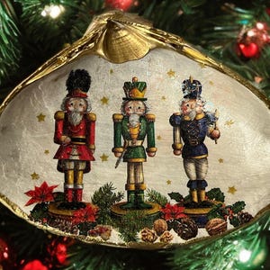 Puede incluir: Un adorno navideño decorativo con tres soldados cascanueces de juguete con uniformes rojos, verdes y azules. La imagen está dentro de una concha dorada, con un tema festivo. El fondo incluye ramas y adornos de árboles de Navidad.