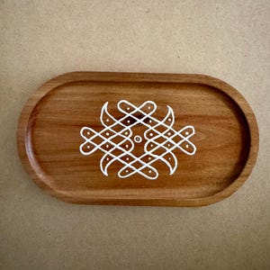 May include: Oval wooden tray with a light brown finish. The tray features a white, symmetrical design in the centre. The design is composed of interconnected lines and dots, creating a decorative pattern. The tray is suitable for serving or display.