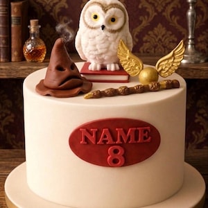 May include: A white cake decorated with a brown Sorting Hat, a white owl, a golden snitch, and a wand. The cake has a red oval with the word "NAME" and the number "8". The cake is on a gold-trimmed base.