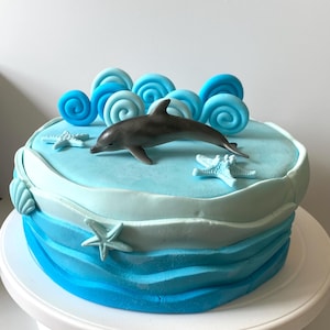 Könnte beinhalten: Eine blaue und weiße Torte, verziert mit einem Delfin, Muscheln und Seesternen. Die Torte steht auf einem weißen Tortenständer.