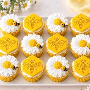 Op de afbeelding: Een set van twaalf cupcakes op een witte schaal, met afwisselende ontwerpen van witte madeliefjes met gele harten en gele zeshoekige plaatjes met een bijenreliëf, elk afgewerkt met een klein fondant bijenfiguurtje.