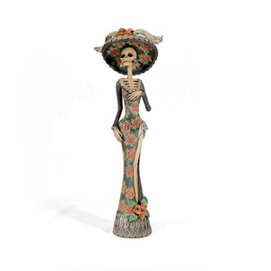 May include: A decorative Day of the Dead skeleton figurine wearing a black dress with orange and green floral patterns and a wide-brimmed hat with orange flowers.