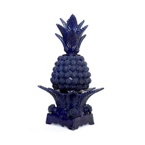 Puede incluir: Una escultura de piña de cerámica azul con un diseño detallado. La piña está sobre una base cuadrada con un diseño de hojas decorativas.
