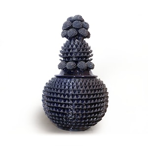 May include: A large, dark blue ceramic vase with a spiky, textured surface. The vase has a round body and a tall, pointed lid with a decorative top.