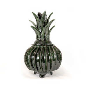 May include: A dark green ceramic pineapple-shaped decorative container with a lid. The container has a ribbed body and a leafy top. The container is on three small feet.