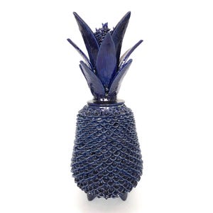 Puede incluir: Un objeto decorativo en forma de piña de color azul oscuro. El objeto tiene una superficie texturizada y una corona de hojas detallada. La piña se apoya sobre tres pequeños pies. El objeto es probablemente de cerámica o vidrio.