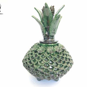 Puede incluir: Un tarro de cerámica verde con forma de piña con una superficie texturizada detallada. El tarro tiene una tapa con una parte superior de piña y una base con tres patas.