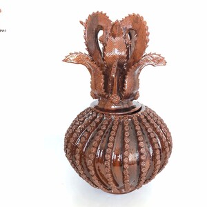 Op de afbeelding: Een bruine keramische vaas met een decoratief deksel. Het deksel is gevormd als een bloem met ingewikkelde details. De vaas is versierd met een patroon van kleine cirkels.