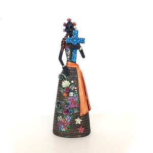 May include: A black skeletal figure with a floral headdress holds a blue cross. The figure is wearing a black and orange dress with floral details. The dress has a black and white pattern and is decorated with colorful flowers.