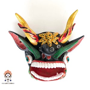 May include: A colorful, hand-painted wooden mask with a black face, red mouth, and white teeth. The mask has green and yellow accents and large, round eyes. The mask is decorated with a yellow and black starburst design on the forehead.