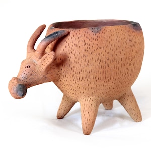 May include: A handcrafted, terracotta-colored planter shaped like a bull. The planter has a textured surface with etched lines, three legs, and curved horns. The bull's snout is a darker shade of brown.