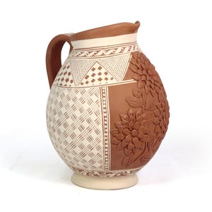 May include: A decorative ceramic pitcher with a handle, featuring a two-tone design. One side has a geometric pattern, the other side has embossed floral details. The pitcher is brown and cream-colored.