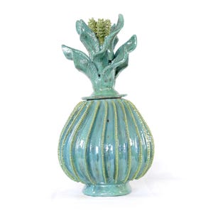 May include: A turquoise ceramic decorative jar with a lid, featuring a unique design. The jar's body has vertical ridges and is topped with a stylized floral element. The overall height is approximately 12 inches.
