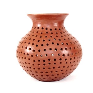 Op de afbeelding: Een terracotta-kleurige keramische vaas met een afgeronde body en een uitlopende rand. De vaas is bedekt met gelijkmatig verdeelde, cirkelvormige gaten. De gaten creëren een decoratief patroon en laten licht door. De vaas wordt waarschijnlijk gebruikt voor decoratieve doeleinden.
