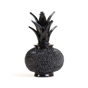Può includere: Un oggetto decorativo nero lucido a forma di ananas. La base presenta una superficie molto strutturata e irregolare, mentre la parte superiore è composta da foglie lisce e appuntite rivolte verso l'alto. L'intero pezzo è di colore nero.