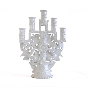 May include: A white ceramic candelabra featuring five candle holders arranged in a tiered structure. The central column and branches are decorated with moulded floral motifs and bird figures, all finished in a glossy, uniform white glaze.