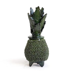 Peut inclure: Un pot en céramique vert foncé en forme d'ananas avec une surface texturée et bosselée, surmonté d'un couvercle amovible aux feuilles pointues. Le récipient repose sur trois petits pieds courts et présente une finition émaillée brillante.