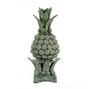 Puede incluir: Una escultura de piña de cerámica verde con una superficie texturizada. La piña tiene una corona detallada y una base con detalles en forma de hojas. La escultura es un elemento decorativo, probablemente para la decoración del hogar.