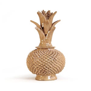 May include: A tan ceramic jar shaped like a pineapple with a textured, bumpy surface and a removable lid featuring sculpted, pointed leaves. The object has a glossy finish and stands on a small, circular base, reflecting light on its surface.