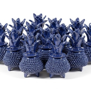 Puede incluir: Colección de recipientes de cerámica azul marino con forma de piña. Cada recipiente tiene un cuerpo texturizado y una corona detallada. Los recipientes están dispuestos en grupo, mostrando su diseño decorativo y forma única. Sobre un fondo blanco.