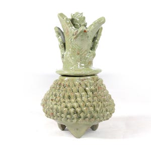 May include: A handmade ceramic pot with a lid, in a light green hue. The body has a textured surface, and the lid has a floral-like design. The pot stands on three small feet. The design is organic and artistic, suitable for decorative use.