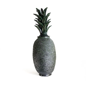 Może przedstawiać: Ciemnozielony ceramiczny pojemnik w kształcie ananasa ze zdejmowaną pokrywką. Korpus posiada teksturowany wzór w kratkę przypominający skórkę ananasa, zwieńczony rzeźbioną, spiczastą zieloną koroną z liści, która służy jako uchwyt pokrywki.