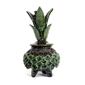 May include: A ceramic decorative object with a dark green and brown color scheme. The object has a pineapple-like design, with a textured, scale-like base and a leafy top. It stands on three small legs.