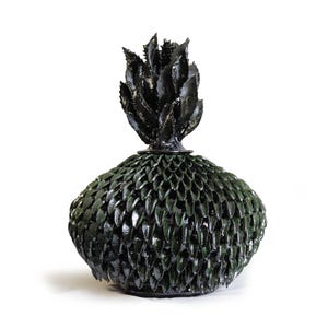 Puede incluir: Un jarrón decorativo de cerámica verde oscuro con un patrón texturizado en forma de escamas en el cuerpo y un diseño espinoso en forma de hojas en la parte superior. El jarrón tiene forma bulbosa y está sobre un fondo blanco.