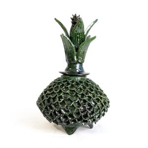 Może przedstawiać: Ciemnozielony, szkliwiony ceramiczny słoik z pokrywką. Korpus posiada teksturowany wzór zachodzących na siebie łusek. Pokrywka ma kształt spiczastej, liściastej korony z ciemnym, teksturowanym środkiem. Całość stoi na trzech małych, zaokrąglonych nóżkach.