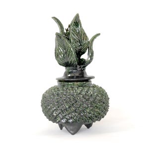 Puede incluir: Escultura de cerámica verde oscuro hecha a mano que representa una planta estilizada. La pieza presenta una base bulbosa texturizada, una sección media lisa y una parte superior con hojas. La escultura está sobre un fondo blanco.