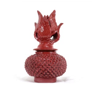 May include: A red ceramic bottle stopper with a textured, round body and a floral-shaped top. The stopper is a deep, glossy red and stands on a white background. The floral top has multiple petal-like elements.