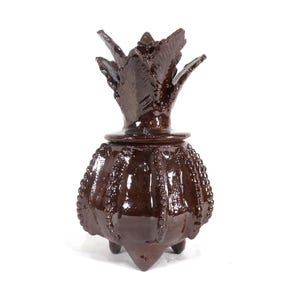 May include: A dark brown decorative jar with a lid, resembling a pineapple. The jar has a textured, ribbed body with a beaded pattern and three pointed feet. The lid is shaped like a crown with pointed leaves.