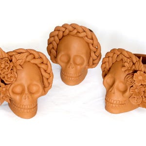 May include: Three terracotta skull-shaped planters. One skull has a braided crown, another has a floral crown, and the third has both. The planters are unglazed and have a matte finish. The image is set against a white background.