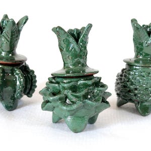 May include: Three dark green ceramic containers with lids. Each has a unique textured base and a stylized, leafy top. The containers are displayed on a white surface, highlighting their intricate details and glossy finish.