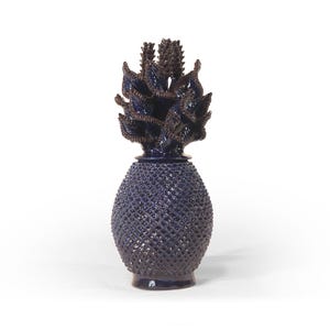 May include: A dark blue ceramic vase with a textured body and a decorative top. The top has a cluster of dark brown, leaf-like elements. The vase is a decorative home accent.