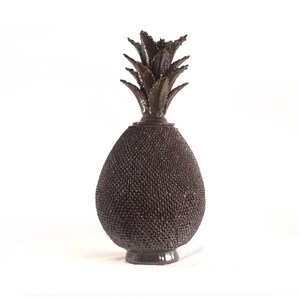 Puede incluir: Un jarrón decorativo en forma de piña, de color marrón oscuro. Tiene una superficie texturizada y una parte superior con hojas detalladas. La base es de un tono marrón más oscuro. Un artículo de decoración único.