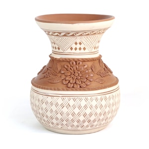 May include: A handcrafted ceramic vase with a cream and brown color scheme. The vase features intricate geometric and floral patterns, including a large flower design around the middle. The vase has a wide base and a flared top.