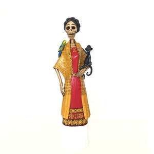 Puede incluir: Una figura de madera pintada a mano de una mujer esqueleto vestida con un vestido amarillo y rojo, sosteniendo un mono negro y un loro verde. La figura es una pieza tradicional de arte popular mexicano.
