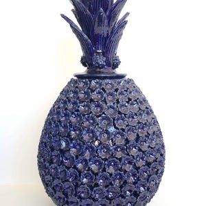 May include: A dark blue ceramic pineapple sculpture. The pineapple's body is covered in small, flower-shaped details. The top has spiky leaves. The sculpture has the text "LA GRAN CATRINA" on the side.