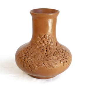 Puede incluir: Un jarrón de arcilla color terracota con cuerpo redondeado y borde acampanado. La superficie exterior presenta un diseño en relieve de tres flores grandes y detalladas similares a crisantemos con pétalos y hojas texturizados.