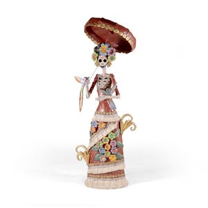 May include: A Day of the Dead figurine depicting a female skeleton in a red dress, holding a bird and a parasol. The dress is decorated with colourful flowers and trim. The figurine has a floral headdress and stands on a white surface.