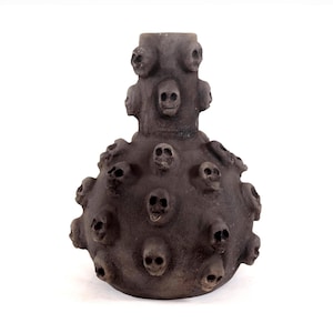 Op de afbeelding: Een donkergrijze decoratieve vaas met een bolvormig lichaam en een cilindrische hals. Het oppervlak is getextureerd en versierd met talrijke kleine schedelvormige gravures, wat een gotische esthetiek creëert. De vaas staat tegen een witte achtergrond.