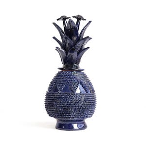 Puede incluir: Un objeto decorativo de cerámica en forma de piña de color azul oscuro con acabado brillante. La base presenta una superficie texturizada con cuentas y un patrón triangular cerca de la parte superior, con una tapa extraíble de hojas puntiagudas.