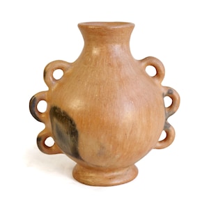 Op de afbeelding: Een terracotta vaas met een rond lichaam, een uitlopende hals en een voetstuk. De vaas heeft aan weerszijden drie symmetrische lusvormige handvatten en vertoont natuurlijke donkere vlekken op het gladde, lichtbruine kleioppervlak.