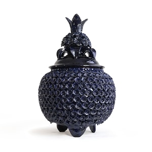 May include: A dark blue ceramic jar with a lid, featuring a dense, textured surface covered in small, raised flower-like shapes. The jar has a rounded body, a pointed finial on the lid, and stands on three short, sturdy feet with a glossy finish.