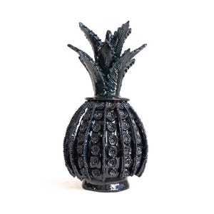 Puede incluir: Un tarro decorativo de cerámica negra brillante con forma de piña y tapa extraíble. El cuerpo presenta crestas verticales adornadas con patrones circulares texturizados, y la parte superior está coronada con hojas puntiagudas y dentadas.