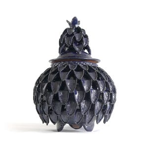 Könnte beinhalten: Dunkelblaue Keramikvase mit Deckel und einzigartigem blattähnlichem Muster. Die Vase hat einen abgerundeten Körper und einen Deckel mit dekorativem Aufsatz. Der Sockel hat drei kleine Füße. Es ist ein dekorativer Artikel.